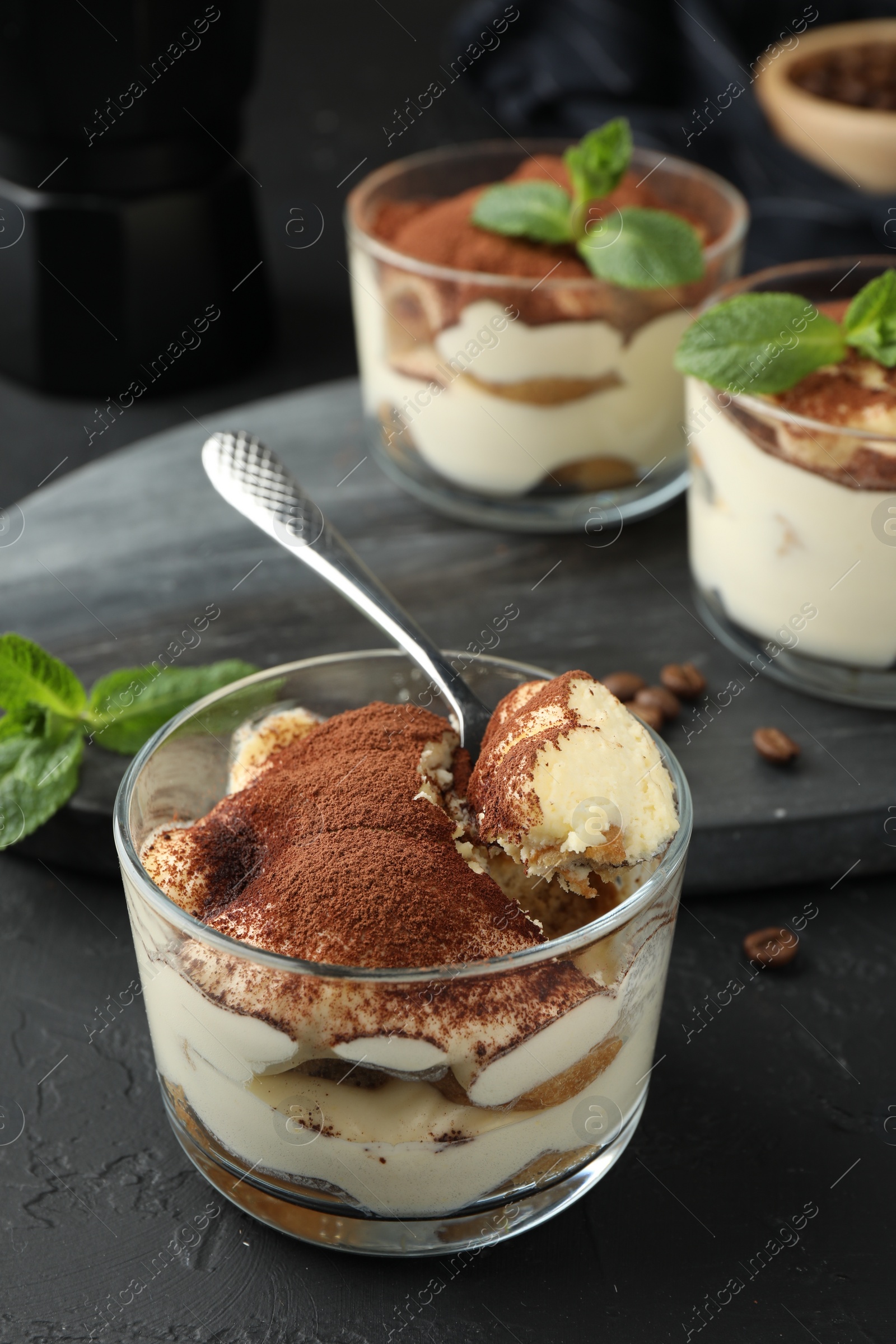 Photo of Delicious tiramisu in glasses and spoon on black table