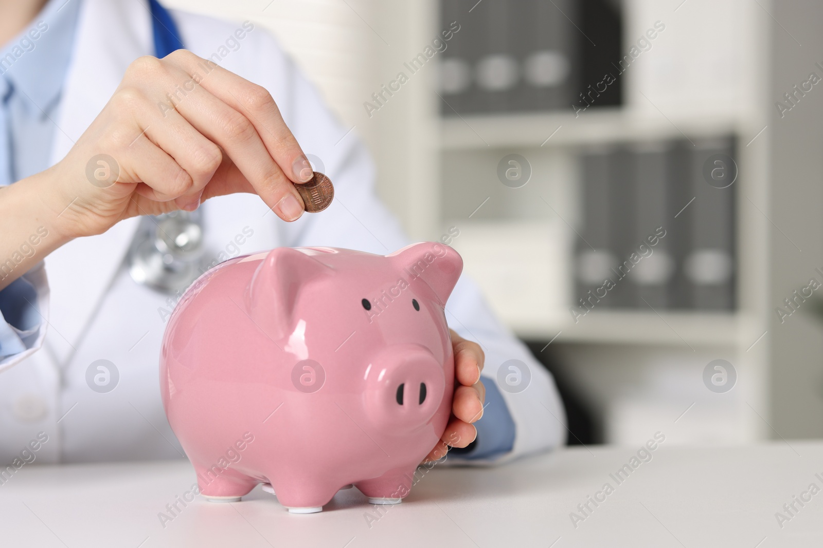 Photo of Doctor putting coin into piggy bank at white table indoors, closeup. Space for text