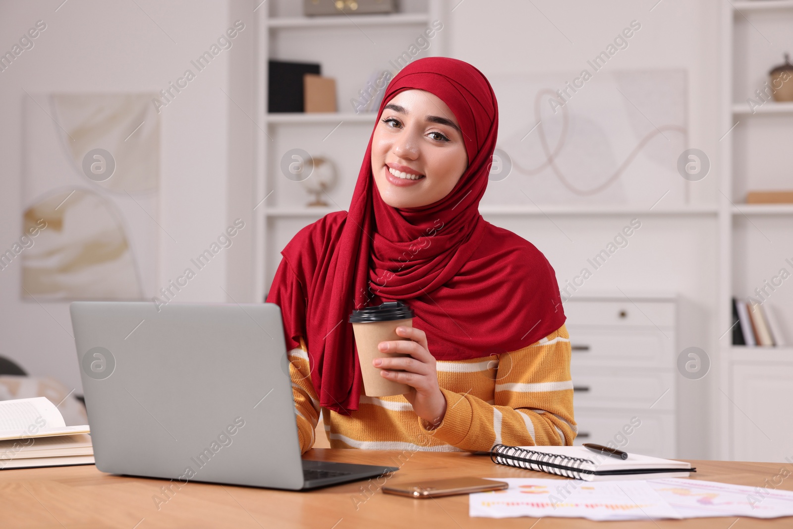 Photo of Muslim woman in hijab with cup of coffee using laptop at wooden table in room