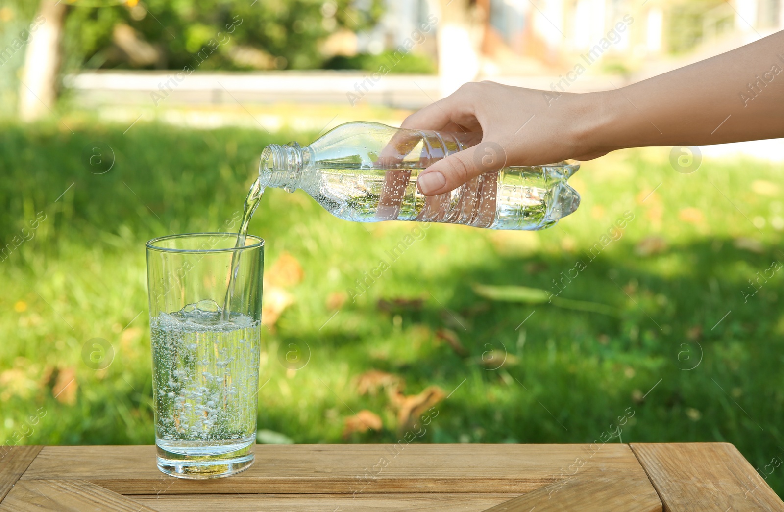 Photo of Woman pouring water from bottle into glass at wooden table outdoors, closeup