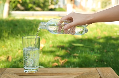 Woman pouring water from bottle into glass at wooden table outdoors, closeup