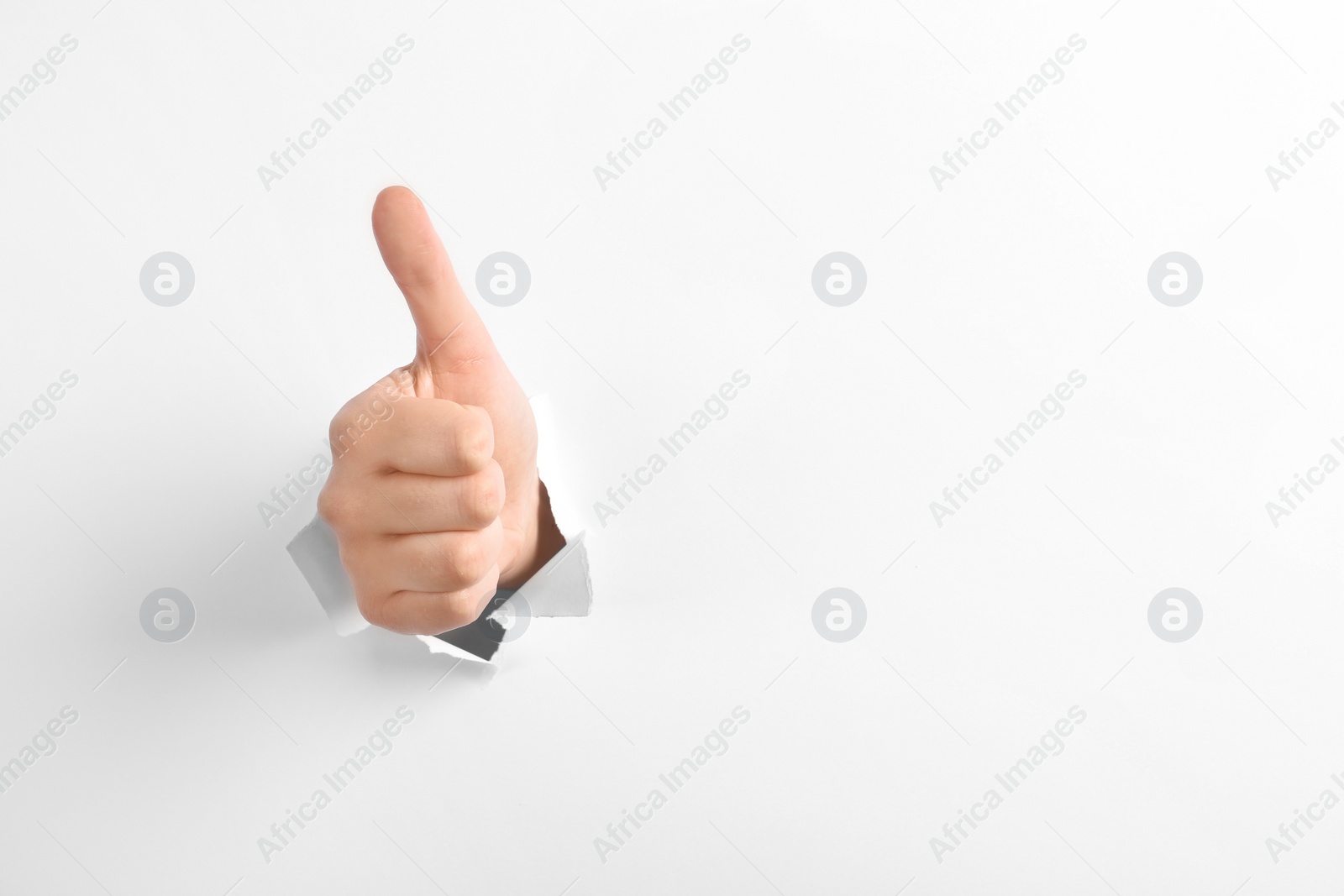 Photo of Woman showing thumbs up gesture through hole in torn white paper, space for text. Victory concept