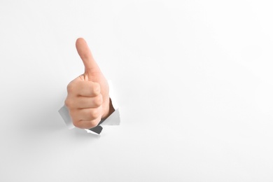 Photo of Woman showing thumbs up gesture through hole in torn white paper, space for text. Victory concept