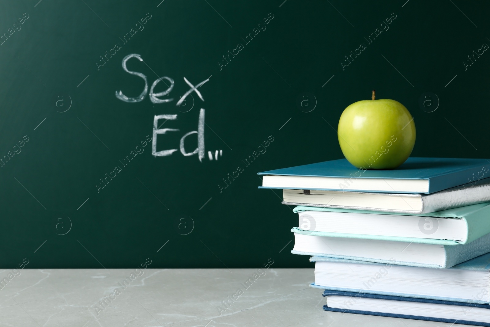 Photo of Books and apple on grey table near chalkboard with phrase "Sex ed"