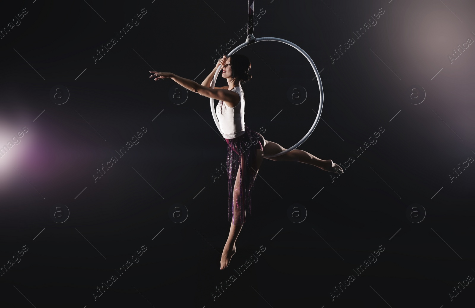 Photo of Young woman performing acrobatic element on aerial ring against dark background