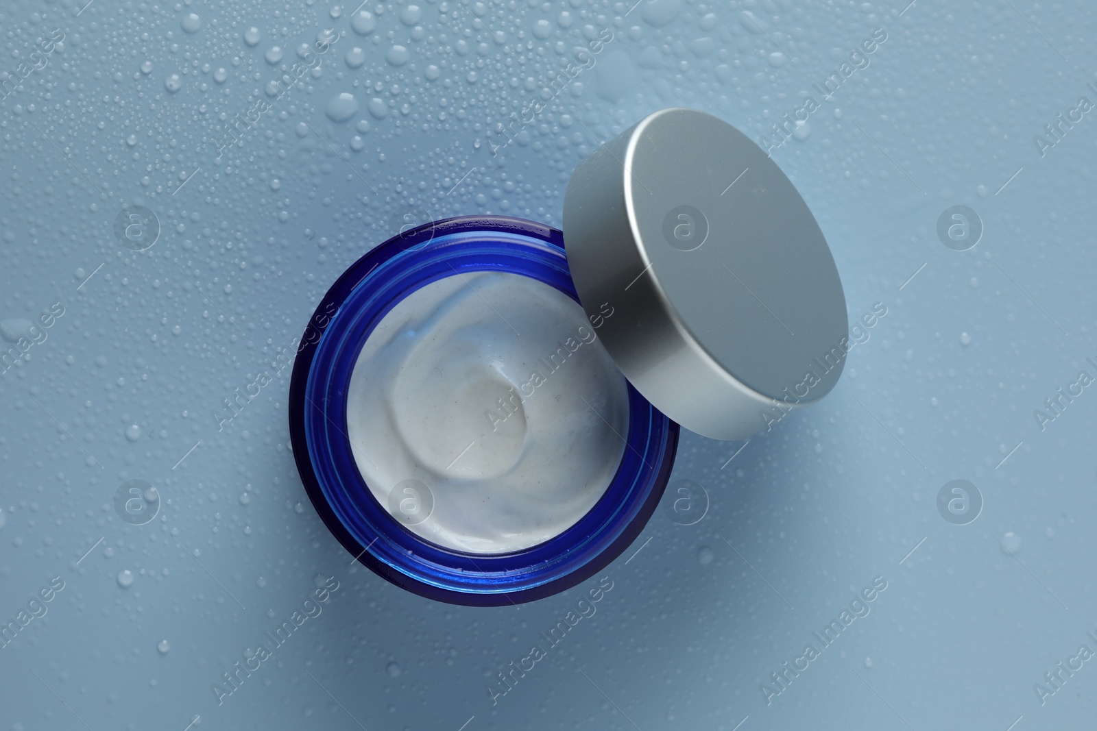 Photo of Moisturizing cream in open jar on light blue background with water drops, top view