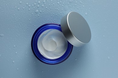 Moisturizing cream in open jar on light blue background with water drops, top view