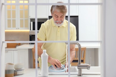 Senior man washing plate above sink in kitchen, view from outside