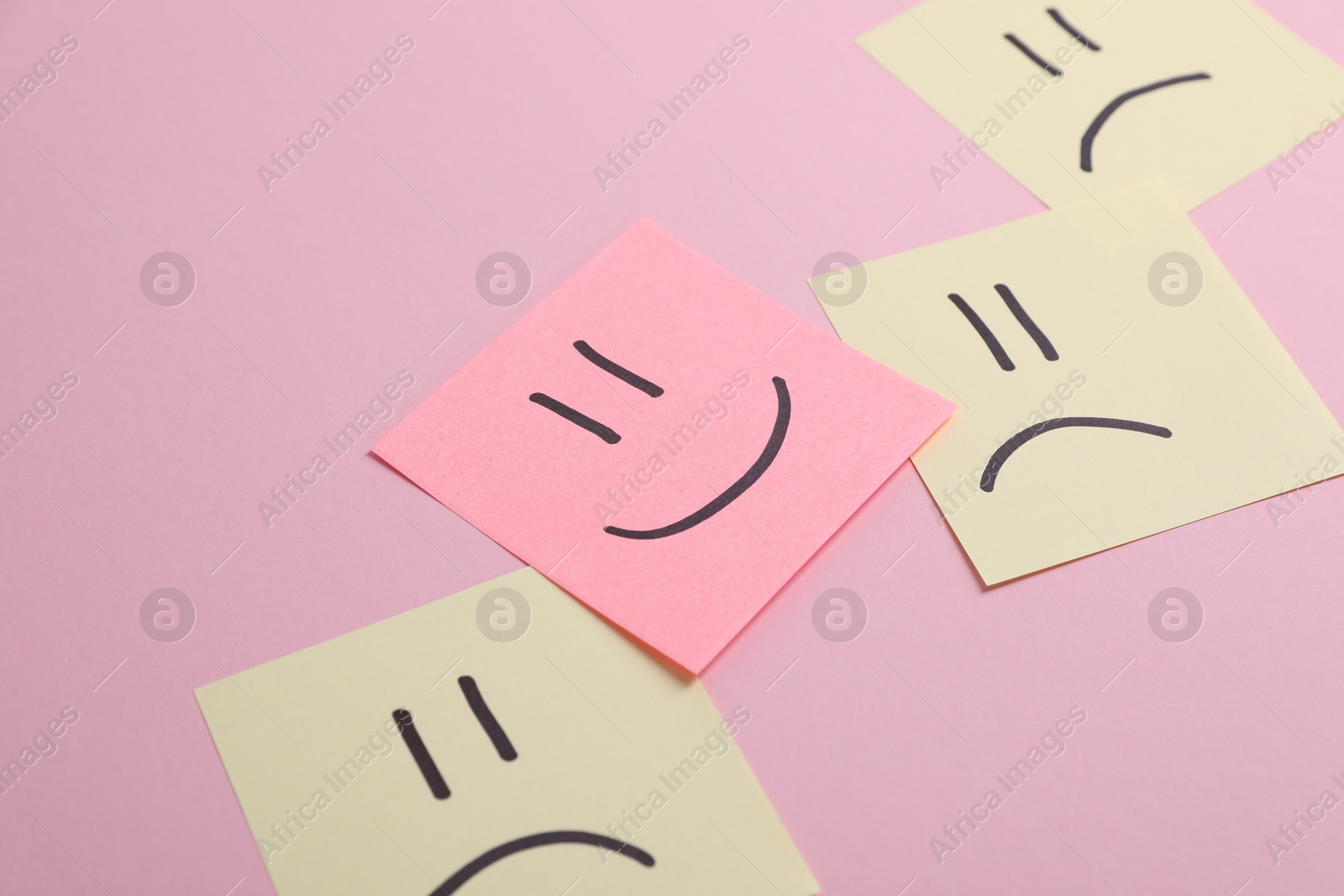 Photo of Choice concept. Sticky note with happy emoticon among beige papers with sad emojis on pink background, closeup