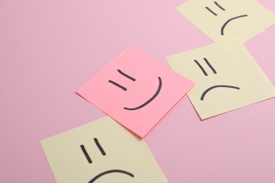 Choice concept. Sticky note with happy emoticon among beige papers with sad emojis on pink background, closeup