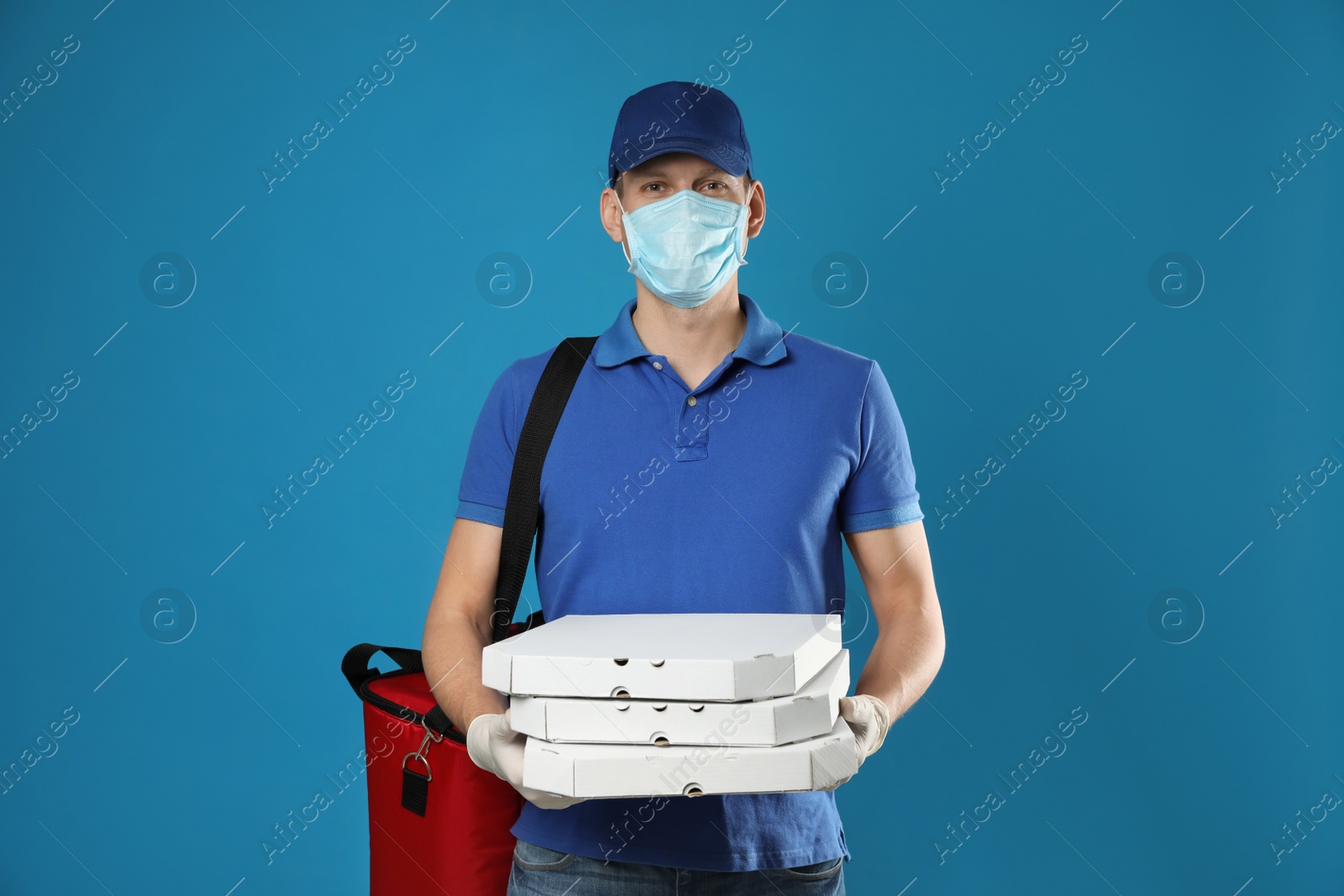 Photo of Courier in protective mask and gloves holding pizza boxes on blue background. Food delivery service during coronavirus quarantine