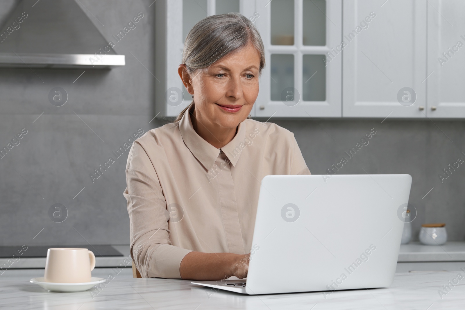 Photo of Beautiful senior woman using laptop at white marble table in kitchen