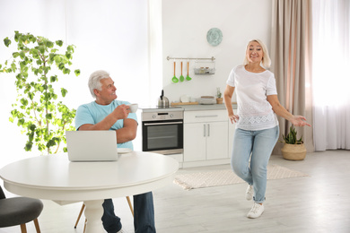 Photo of Middle aged woman dancing near her husband in kitchen. Happy mature couple