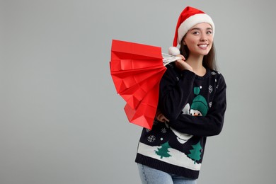Photo of Young woman in Christmas sweater and Santa hat with shopping bags on grey background. Space for text