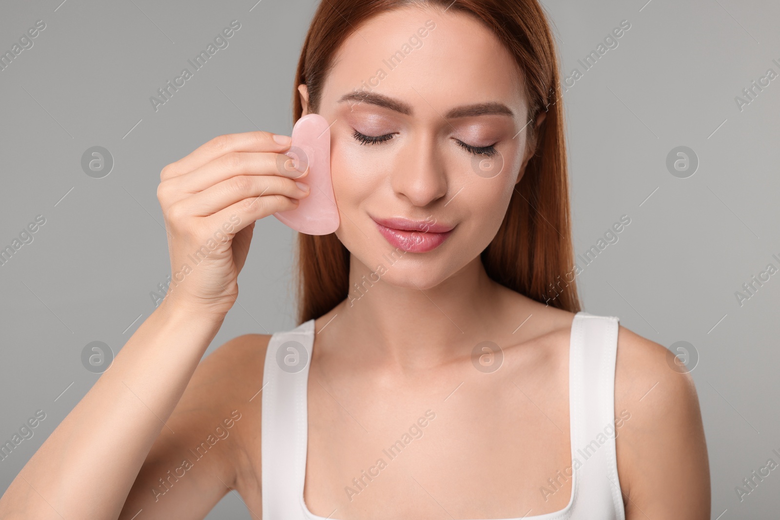 Photo of Young woman massaging her face with rose quartz gua sha tool on grey background