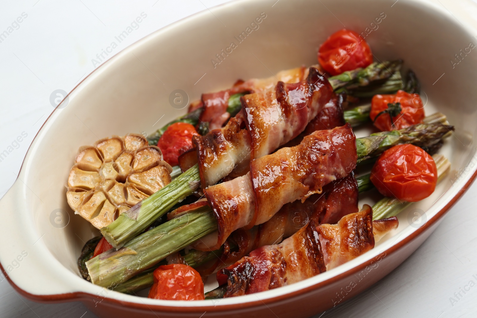 Photo of Oven baked asparagus wrapped with bacon in ceramic dish on white table, closeup