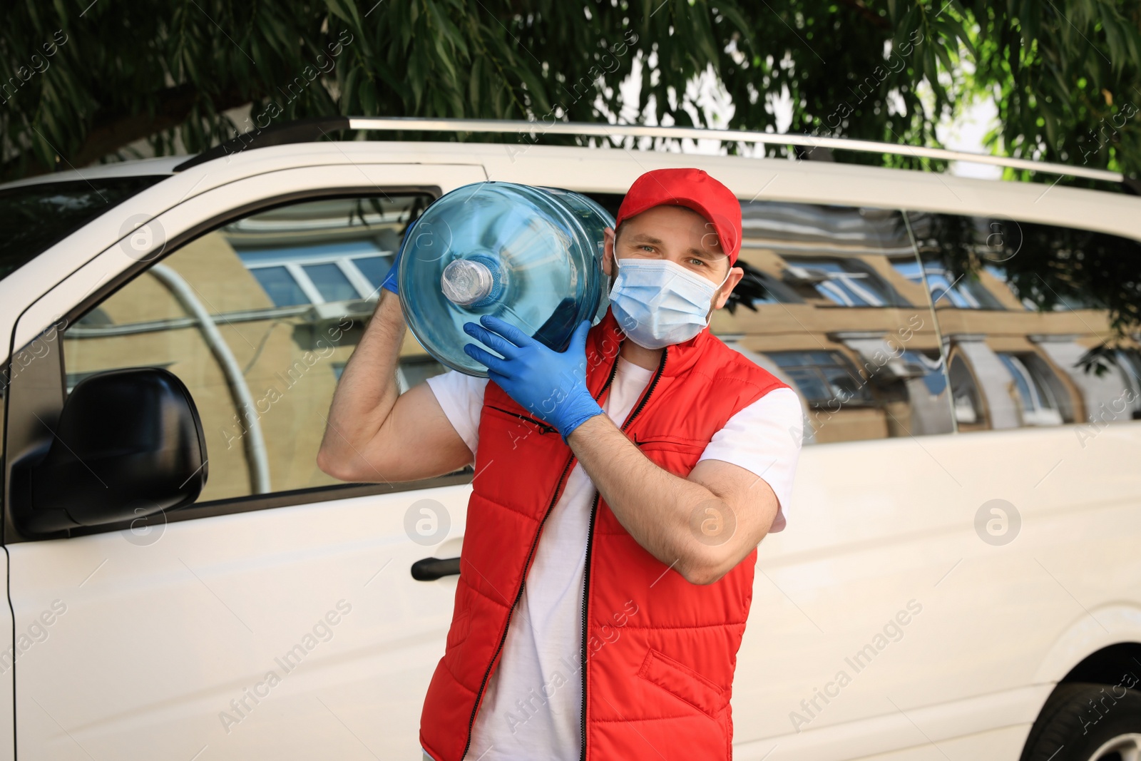 Photo of Courier in medical mask holding bottle of cooler water near car outdoors. Delivery during coronavirus quarantine