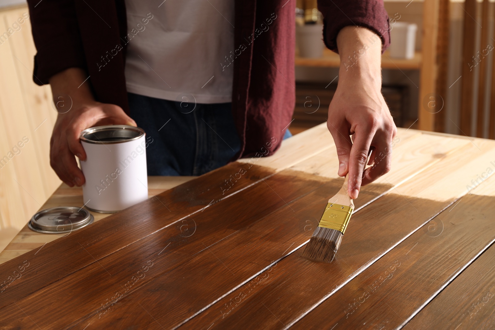 Photo of Man with brush applying wood stain onto wooden surface indoors, closeup