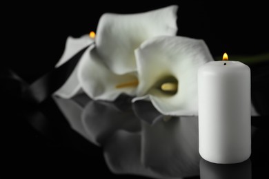 Photo of Burning candle and white calla lily flowers on black mirror surface in darkness, closeup with space for text. Funeral symbol