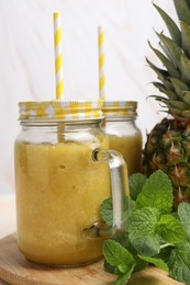 Photo of Tasty pineapple smoothie in mason jars, mint and fruit on wooden board, closeup