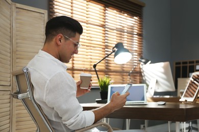 Photo of Freelancer with cup of coffee using smartphone while working indoors. Space for text