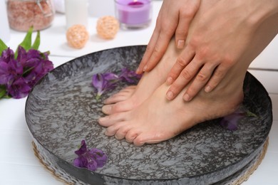 Photo of Woman soaking her feet in bowl with water and flowers on white floor, closeup. Spa treatment