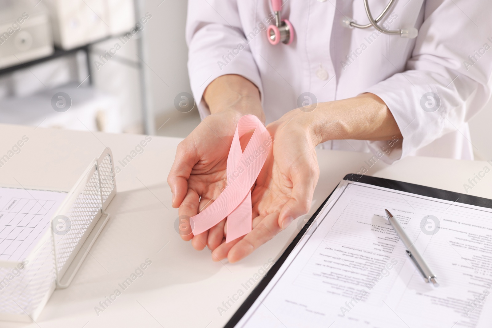 Photo of Doctor holding pink ribbon at white desk indoors, closeup. Breast cancer awareness