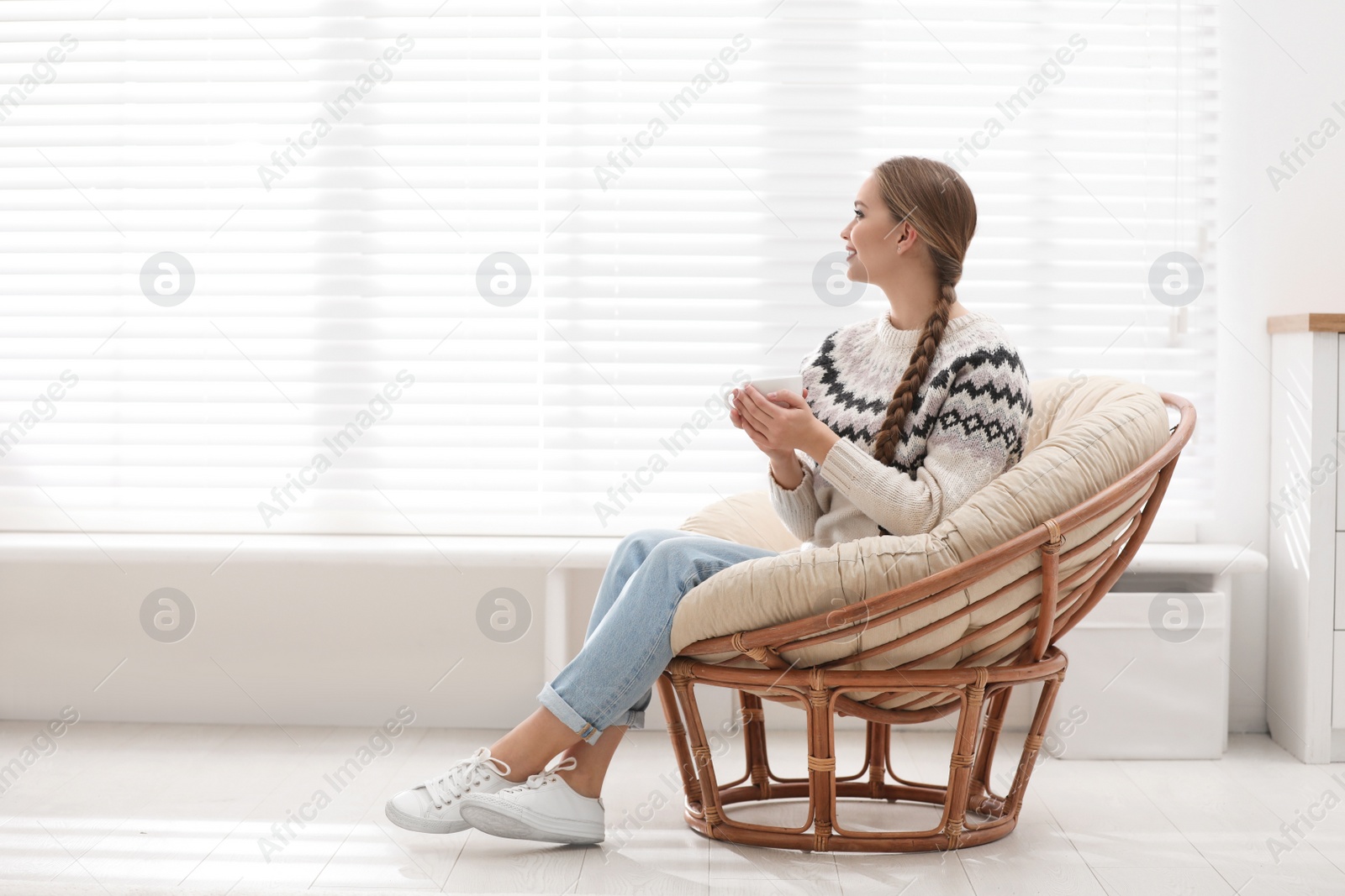 Photo of Young woman with hot drink in papasan chair near window at home