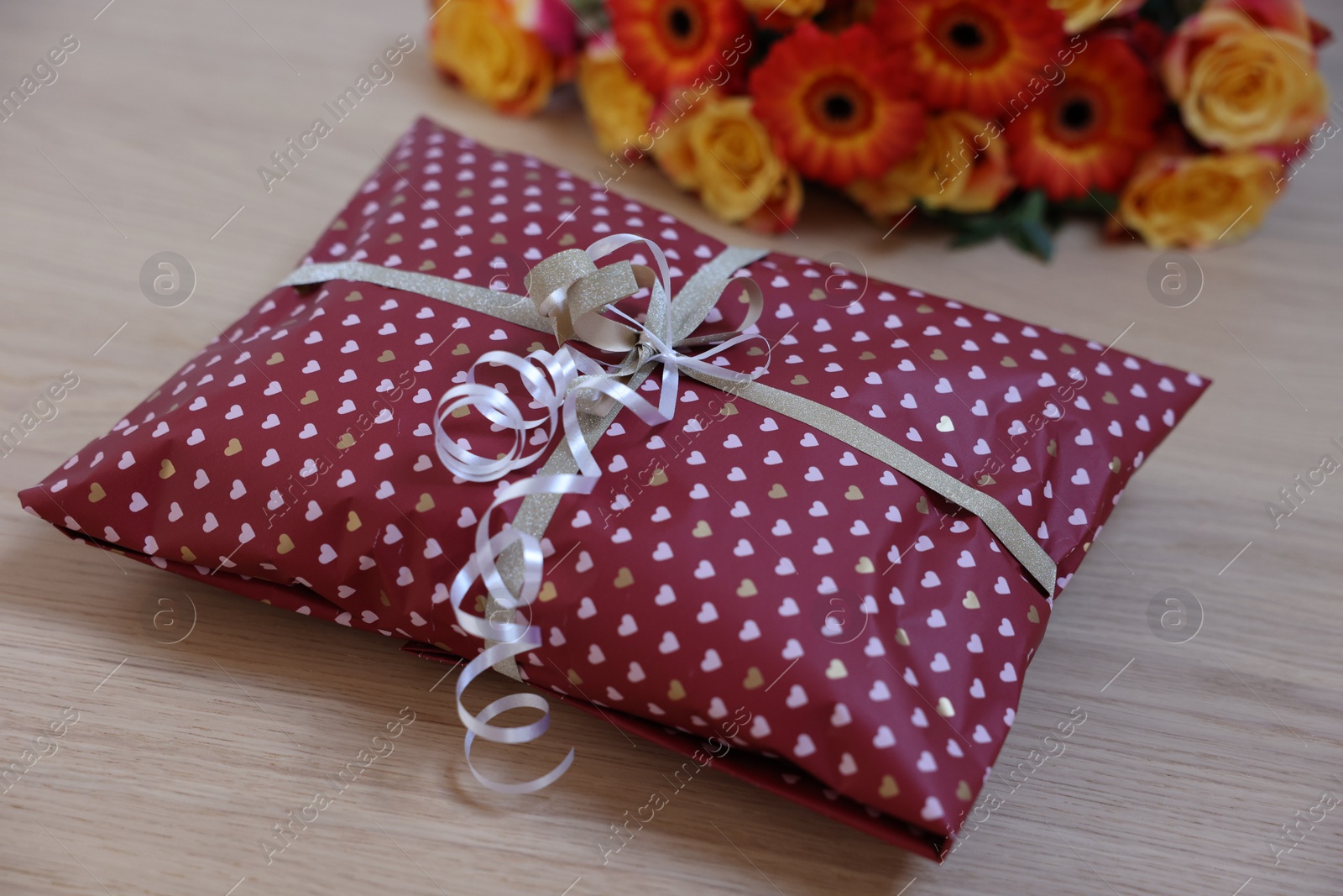 Photo of Parcel wrapped in heart patterned paper and beautiful flowers on wooden table, closeup