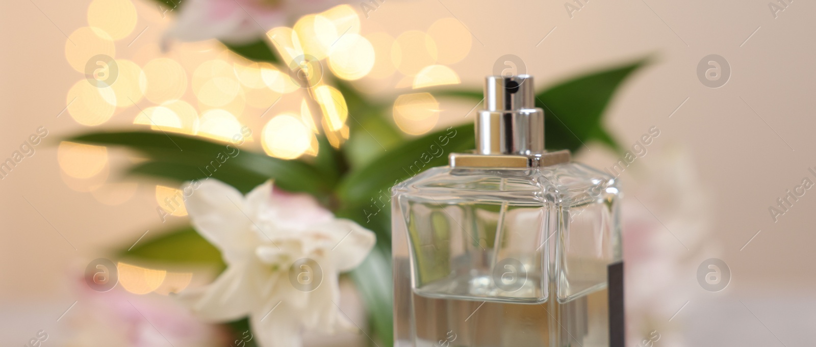 Photo of Bottle of perfume and beautiful lily flowers against beige background with blurred lights, closeup