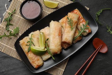 Tasty fried spring rolls, arugula, lime and sauce on dark textured table, flat lay