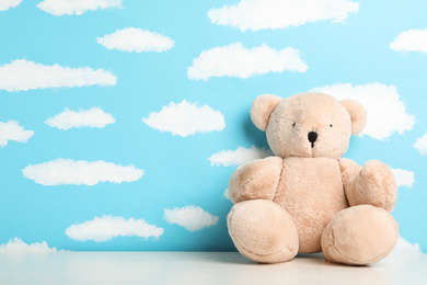 Photo of Teddy bear on white table near wall with painted blue sky, space for text. Baby room interior