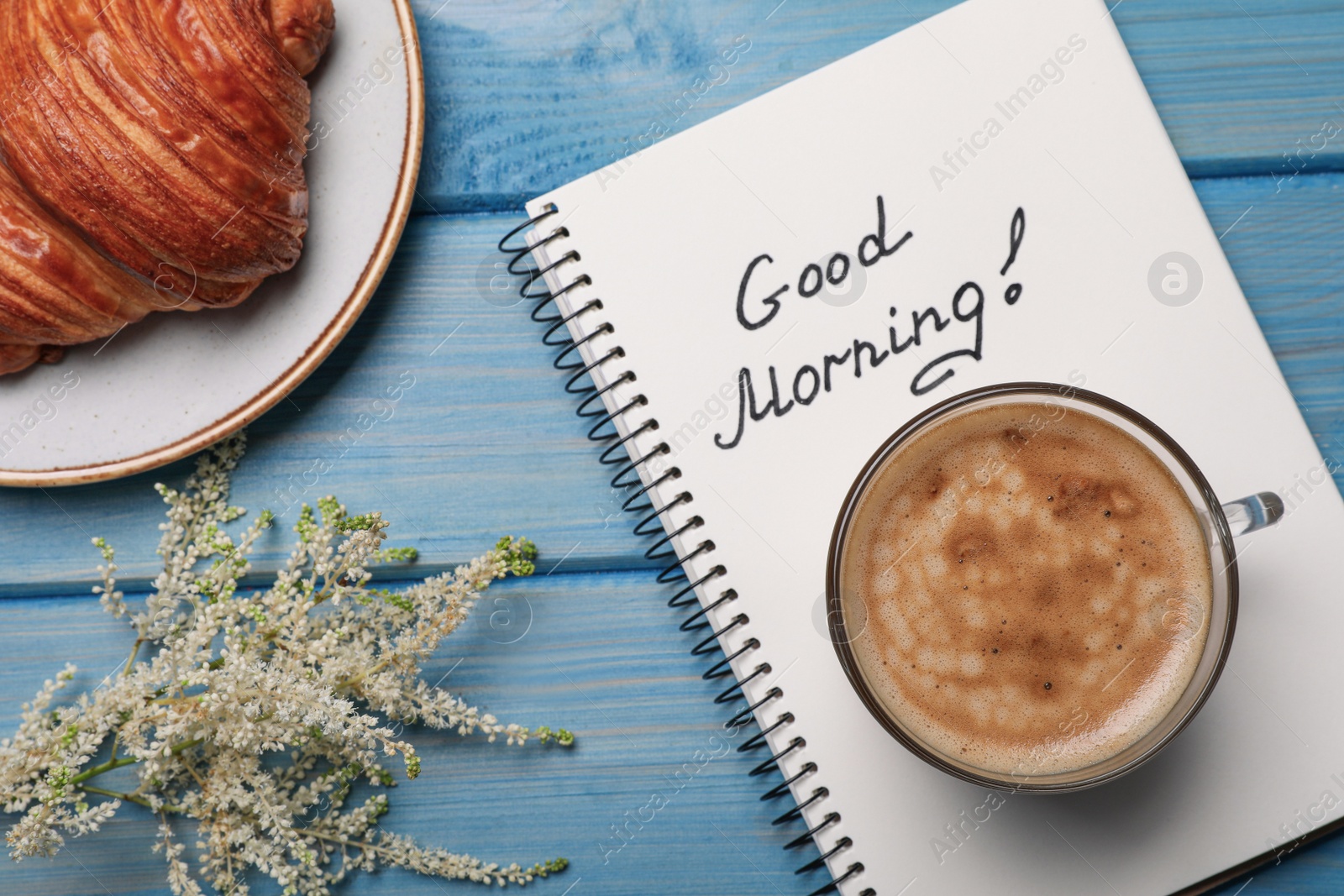 Photo of Aromatic coffee, croissant, flowers and Good Morning! message on light blue wooden table, flat lay