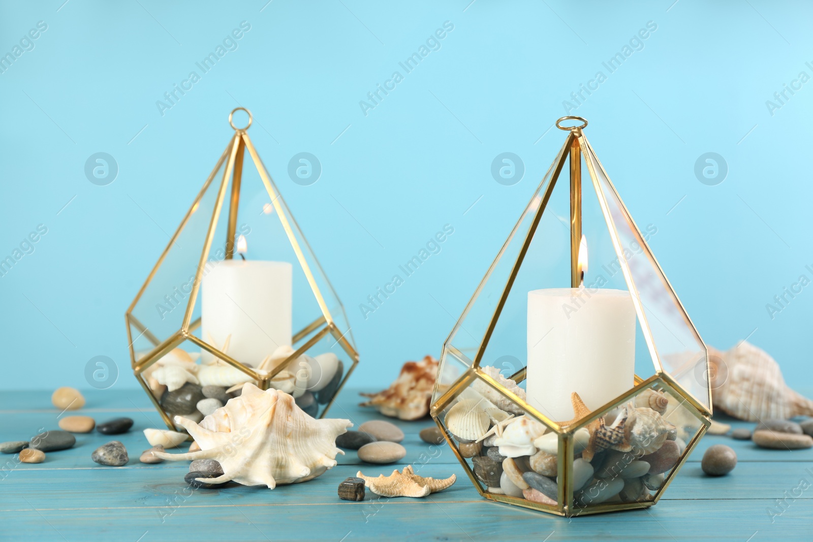 Photo of Stylish glass holders with burning candles, seashells and pebbles on light blue wooden table