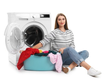 Photo of Young woman sitting near washing machine with basket of laundry on white background