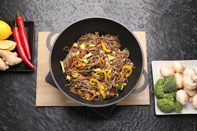 Photo of Stir-fry. Tasty noodles with vegetables, meat in wok and ingredients on dark textured table, flat lay