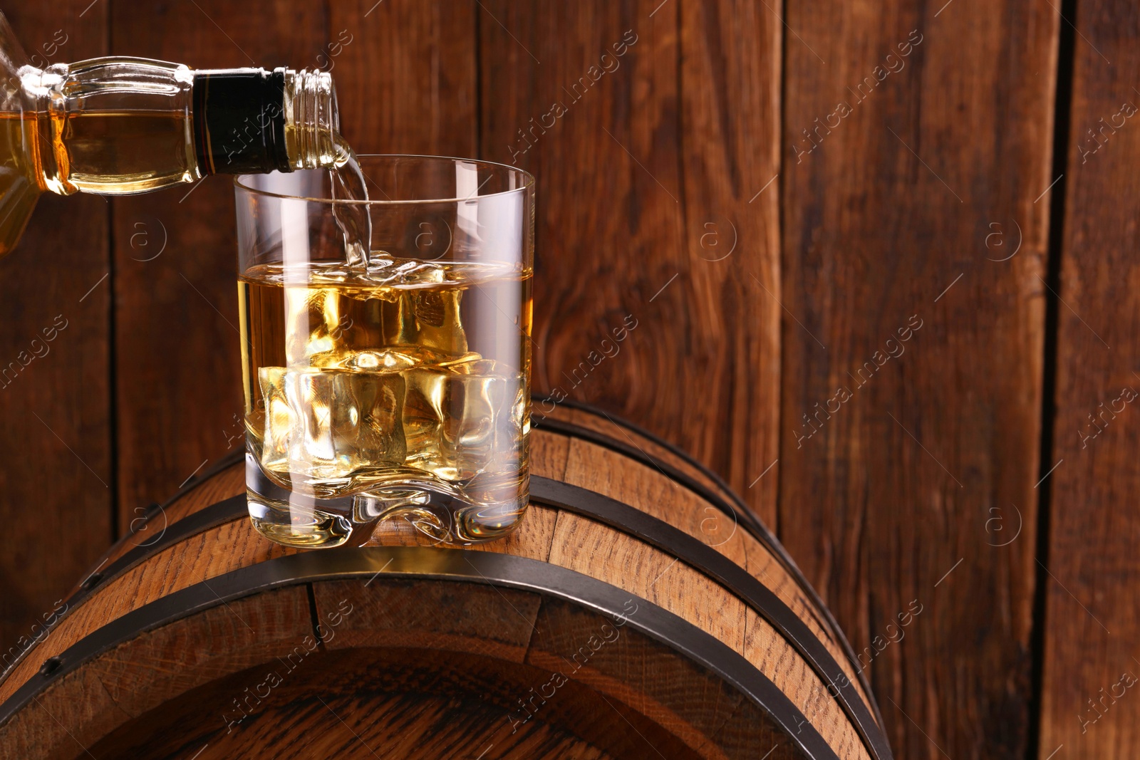 Photo of Pouring whiskey from bottle into glass on barrel against wooden background, closeup. Space for text