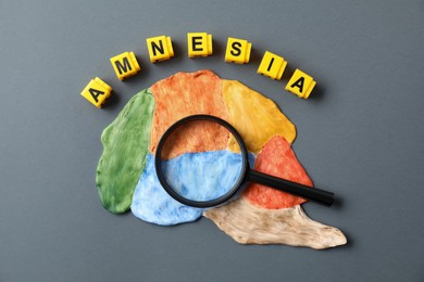 Word Amnesia made of yellow cubes, magnifying glass and brain with sections on grey background, flat lay