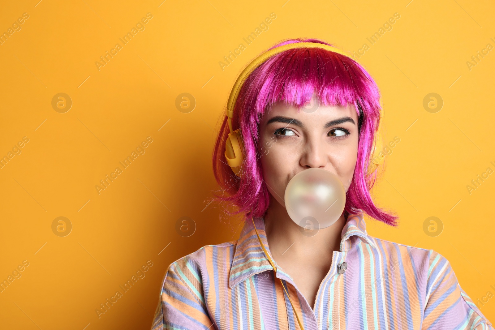 Photo of Fashionable young woman in colorful wig with headphones blowing bubblegum on yellow background, space for text
