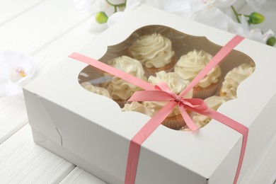 Tasty cupcakes with vanilla cream in box and orchid flowers on white wooden table, closeup