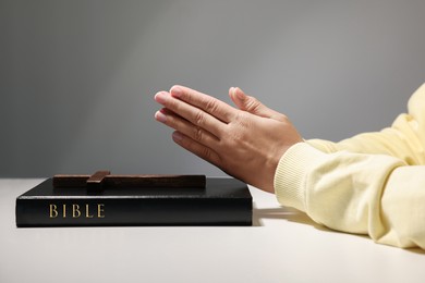Photo of Woman praying over Bible with wooden cross at white table against grey background, closeup