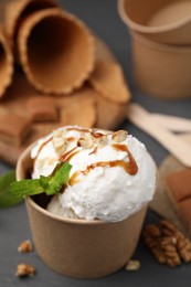 Photo of Tasty ice cream with caramel sauce, mint and nuts in paper cup on grey wooden table