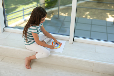 Photo of Little girl drawing rainbow near window indoors. Stay at home concept