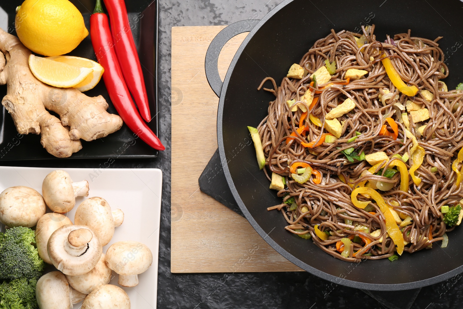 Photo of Stir-fry. Tasty noodles with vegetables, meat in wok and ingredients on dark textured table, flat lay