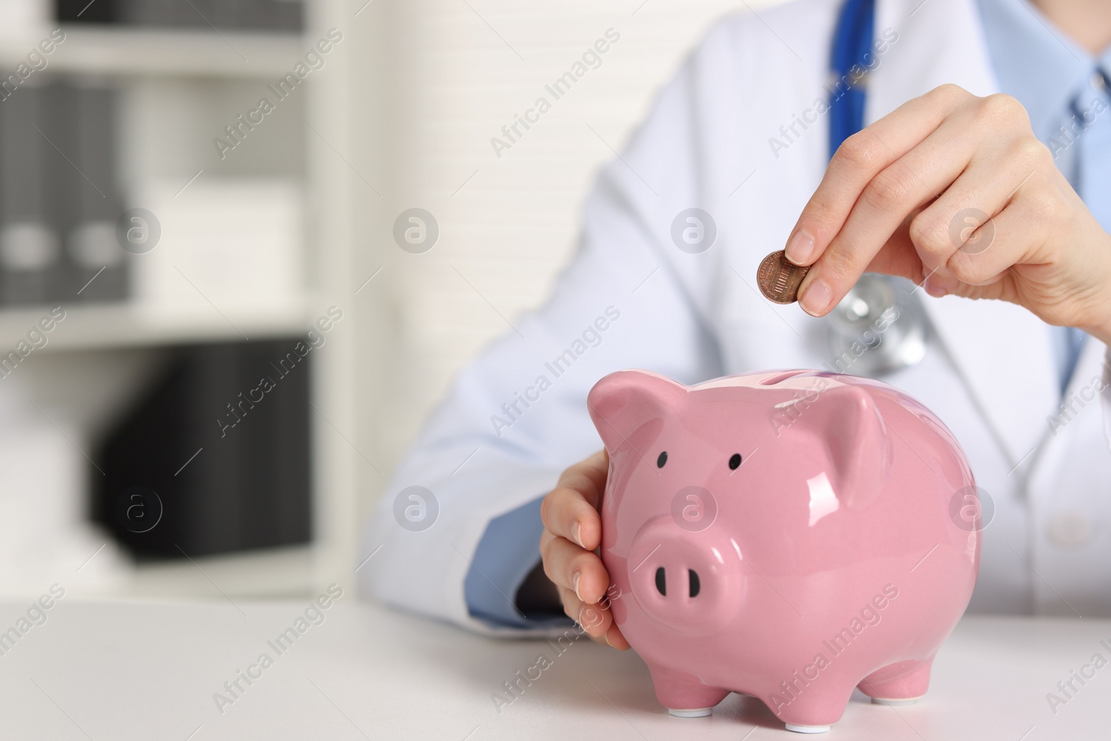 Photo of Doctor putting coin into piggy bank at white table indoors, closeup. Space for text