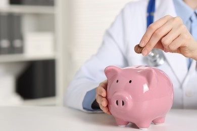 Photo of Doctor putting coin into piggy bank at white table indoors, closeup. Space for text