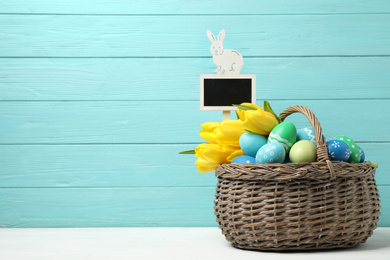 Colorful Easter eggs with fresh tulips and tag in wicker basket on white table against light blue background. Space for text