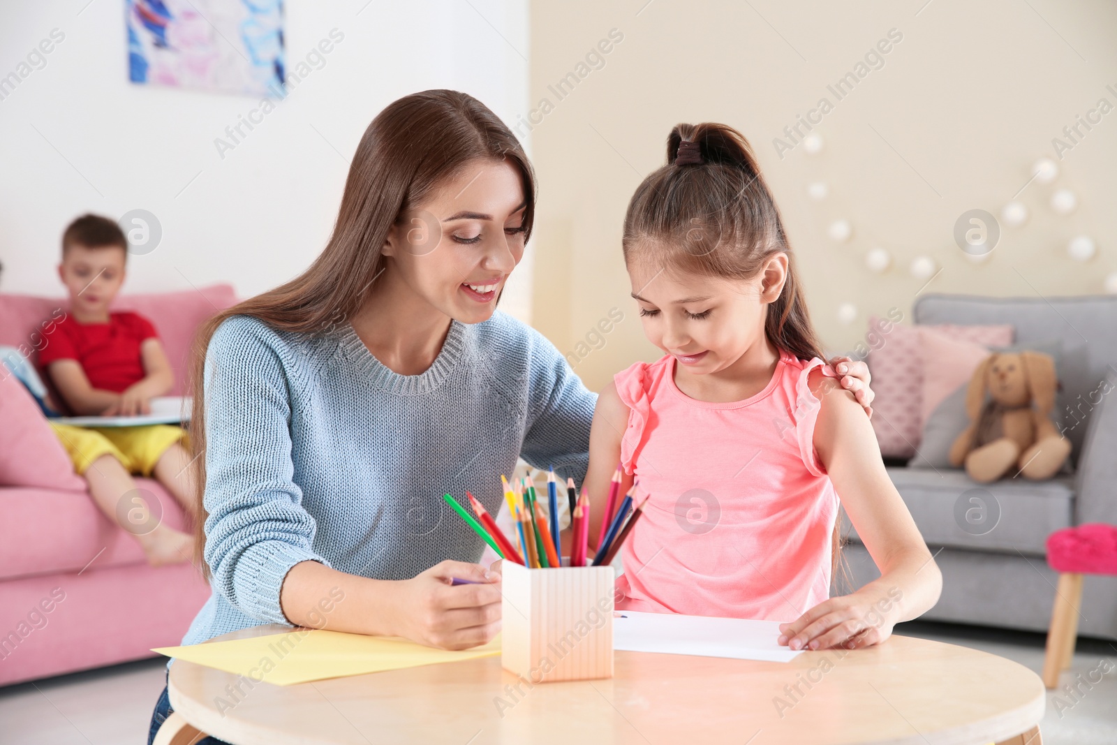 Photo of Cute little child drawing at table with young woman in playing room