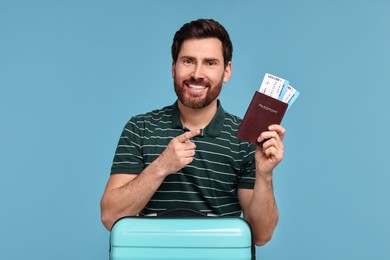 Smiling man pointing at passport and tickets on light blue background