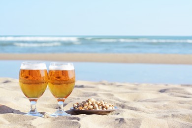 Photo of Glasses of cold beer and pistachios on sandy beach near sea, space for text